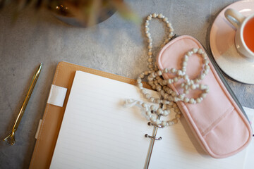Opened notebook with clean sheets, gold pen, rosary, pink colored case and cup of tea near vase