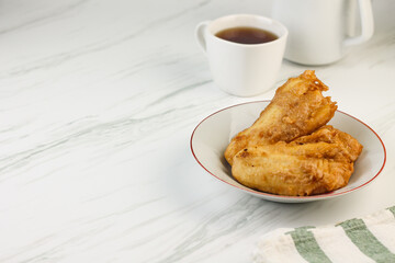 Pisang Goreng or Banana fritters, Indonesian traditional snack made from banana and flour, deep fried. Served on white plate, with a cup of tea. Isolated white background. Copy space for text. 