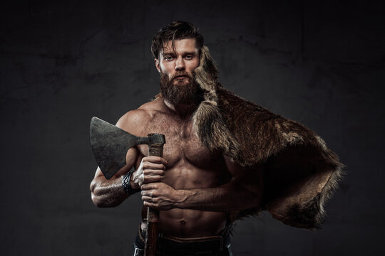 Medieval scandinavian warrior with muscular naked build posing with his hunting trophy and axe in dark studio background.