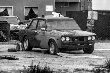 Black and white image of a rusty, old, abandoned car in an empty dirt lot full of weeds.