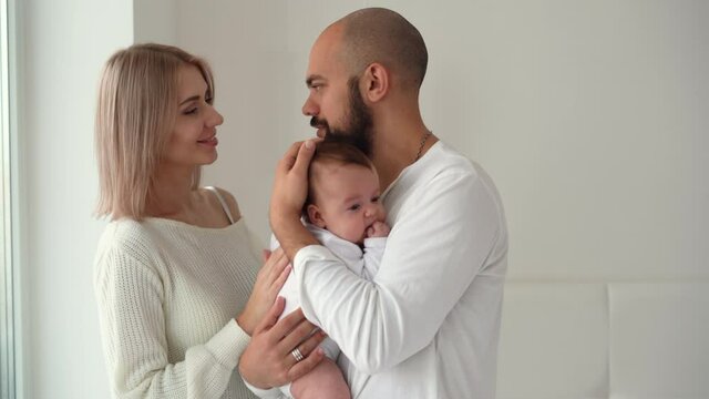 Mother, father and his newborn baby standing in the room. Happy parents kiss and hug the newborn. Paternity concept. Motherhood Beautiful happy young family.