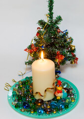 Burning Christmas candle with red balls, a toy rooster, glass beads on the plate and Christmas tree
