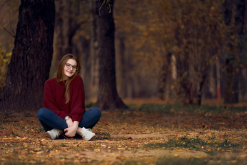Woman with freckles in glasses with crossed legs sits on nature. Autumn forest.