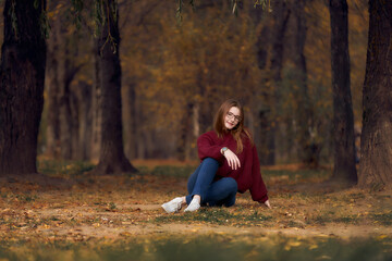 A woman with glasses and a burgundy sweater with red hair sits on a forest path. Beautiful pine autumn forest.