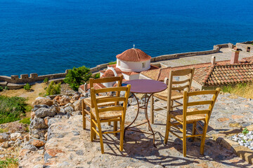 Traditional cafe exterior in the fortified medieval  castle of Monemvasia. Iron tables and wooden chairs with the view of the church panagia chrysafitissa and the aegean sea in the background.