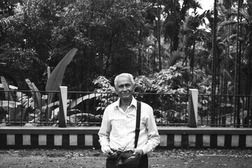 An old man, wearing spectacles and shirt, standing alone in Mawlynnong village, East Khasi Hills, Meghalaya, India. The elder person with cap in hand, side bag on shoulder, looking towards the camera.