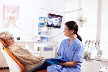 Dental assistant completing questioner during oral checkup of elderly patient. Senior woman talking with medical nurse in stomatology office about teeth problem.