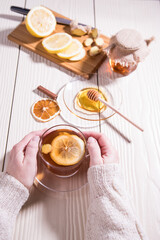 Women's hands in a white knitted sweater hold a Cup of vitamin tea with lemon, honey and ginger, white background.Close up. Vertical orientation.