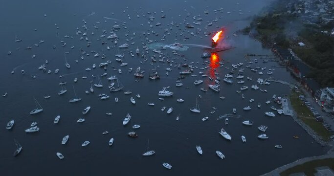 Extremely interesting, high altitude, static aerial footage of small motor boats, sail boats gathering around and witnessing the biggest bonfire event on Earth at Alesund, Norway at dusk.