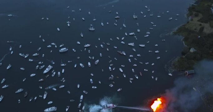 Extremely interesting, high altitude, upward revealing aerial footage of small motor boats, sail boats gathering around and witnessing the biggest bonfire event on Earth at Alesund, Norway at dusk.