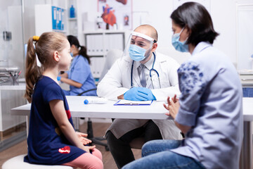 Fototapeta premium Physician having conversation with child during consultation and coronavirus outbreak in hospital office wearing face mask. Health pediatrician specialist providing health care services consultations