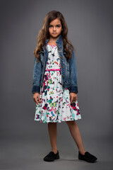 charming little girl in dress and denim jacket posing on gray background
