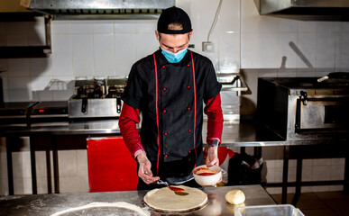 
Pizza cooking concept, chef in medical mask against covid-19 puts ketchup on pizza