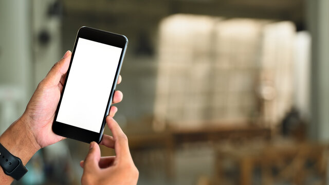 Mockup image of hands holding  smart phone with blank white screen and living room bluer background.