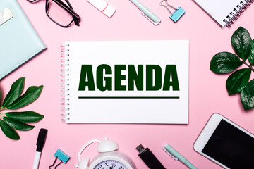 AGENDA is written in a white notebook on a pink background surrounded by business accessories and green leaves.