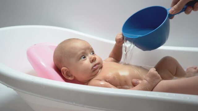 A hand pours water on a baby in the bathroom from a blue bucket
