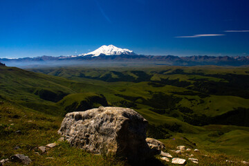 An excellent view from the plateau to the extinct volcano and the snowy peak of Mount Elbrus. Below is a green gorge. Stone in the foreground