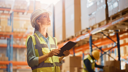 Professional Female Worker Wearing Hard Hat Checks Stock and Inventory with Digital Tablet Computer in the Retail Warehouse full of Shelves with Goods. Working in Logistics, Distribution Center 