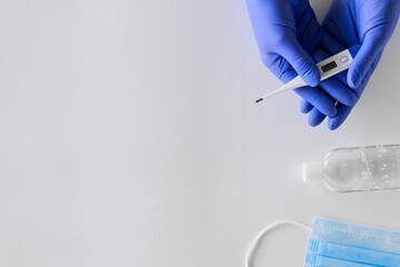 female hands in blue gloves hold a white thermometer, an antiseptic and a blue mask lies on a light background