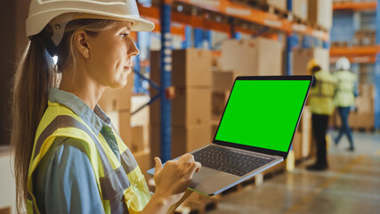 Professional Female Worker Wearing Hard Hat Holding Laptop Computer with Green Chroma Key Screen in Landscape Mode in the Retail Warehouse full of Shelves with Goods. Over the Shoulder Side view 