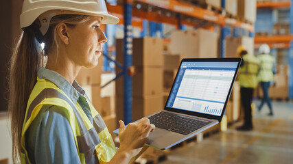 Professional Female Worker Wearing Hard Hat Holds Laptop Computer with Screen Showing Inventory Checking Software in the Retail Warehouse full of Shelves with Goods. Over the Shoulder Side View 