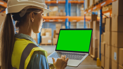 Professional Female Worker Wearing Hard Hat Holding Laptop Computer with Green Chroma Key Screen in Landscape Mode in the Retail Warehouse full of Shelves with Goods. Over the Shoulder Side view 