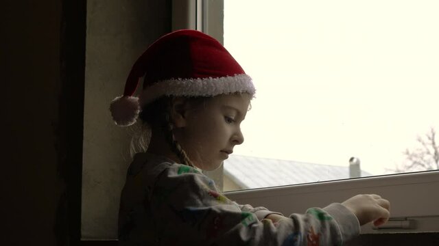 Little girl by the window in Santa hat. Orphans and Christmas . Orphanage life and holidays.