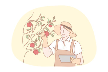 Farming, agriculture, harvesting concept. Smiling young man farmer in working apron, gloves and hat standing with documents and touching fresh red ripe apples on tree in farmland for harvesting 