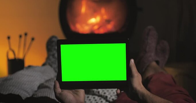 Relaxed couple rest on the sofa in front of fireplace watch green screen tablet wearing woollen socks,winter christmas scene,holding chroma key screen display by the fire place