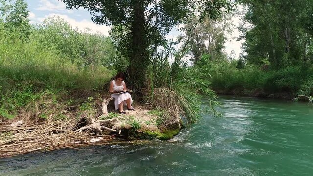 Adolescente leyendo libro bajo la sombra de un &aacute;rbol junto a un r&iacute;o bravo y dron orbitando sobre ella