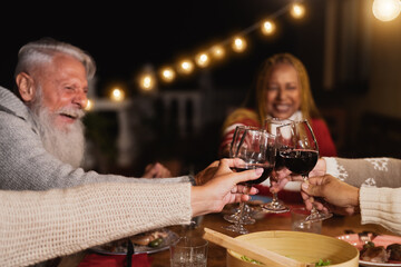 Happy multiracial seniors toasting with red wine glasses and celebrating holidays on house patio dinner - Elderly lifestyle people concept