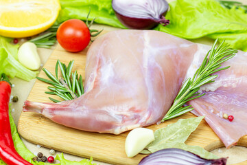 Raw uncooked pink rabbit leg meat with green rosemary and fresh vegetables on light wooden cutting board background.