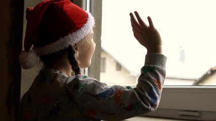 Fototapeta na wymiar Little girl by the window in Santa hat. Orphans and Christmas . Orphanage life and holidays.