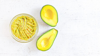 Two avocado halves, and glass bowl with prepared guacamole on white stone like desk, view from above space for text right side