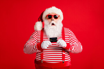 Portrait of his he nice handsome amazed stunned wondered bearded fat Santa holding in hand bank card modern novelty service reaction isolated over bright vivid shine vibrant red color background