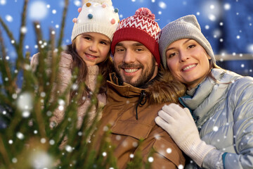Fototapeta premium family, winter holidays and people concept - happy mother, father and little daughter choosing christmas tree at street market in evening over snow