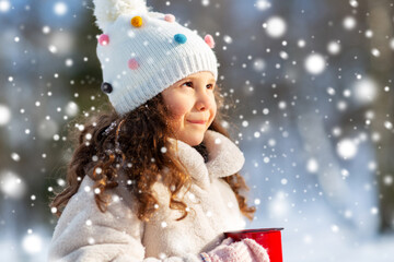 childhood, christmas and season concept - happy little girl with cup of hot tea in winter park over snow