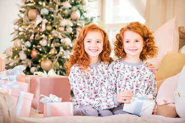 Red-haired children in pajamas sitting on bed with gifts in their hands on the background of the Christmas tree.