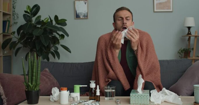 Adult ill guy talking paper handkerchief and sneezing while sitting on sofa covered with blanket.Young bearded man feeling bad while having flu or cold and taking cure at home