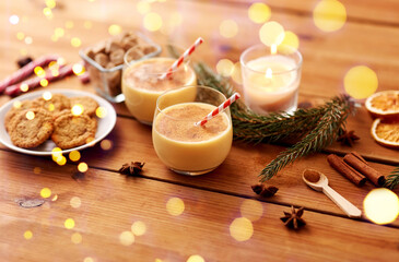 christmas and seasonal drinks concept - glasses of eggnog with oatmeal cookies, candy canes, sugar, fir tree branches and candle burning on wooden background over lights
