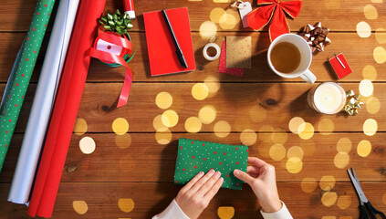 holidays, new year and christmas concept - hands wrapping gift box into green paper on wooden table over lights
