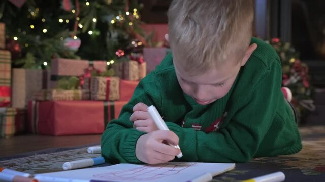 Small blond boy in green sweater lies on floor near Christmas tree and writes letter with markers on paper to Santa Claus. Child writes letter with wishes for gift for Santa Claus.