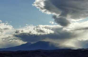 Cime montuose coperte da grandi nuvole bianche e raggi di sole  nel cielo azzurro di in un tramonto invernale