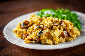 scrambled eggs with mushrooms and arugula salad in white plate on wooden table background