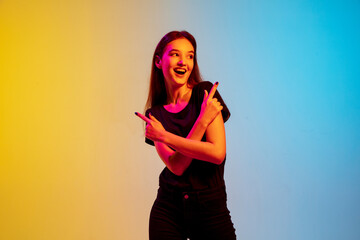 Pointing, laughting. Young caucasian woman's portrait on gradient blue-yellow studio background in neon light. Concept of youth, human emotions, facial expression, sales, ad. Beautiful brunette model.