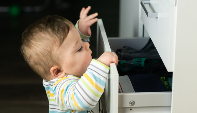 Little Caucasian Baby Boy Opening A White Drawer With Underwear When Nobody Is Watching. Naughty Toddler Exploring Dangerous Furniture. Baby Safe At Home Concept.