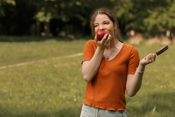 Fototapeta premium Close up lifestyle portrait of young girl college student devouring juicy red apple and holding ice lolly cream in hand. Woman in city park in sunny day. Image with blur background, copy empty space.