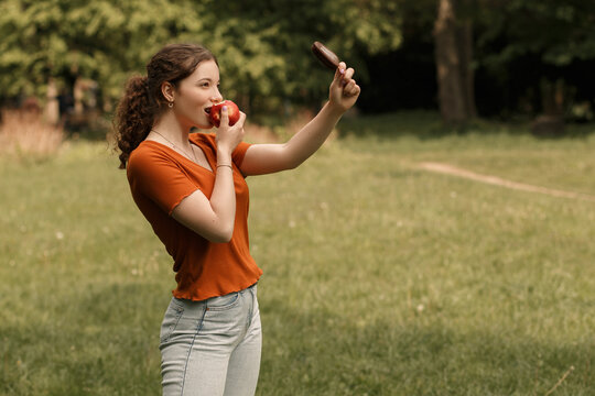 Lifestyle Portrait Of Smiling Curly Girl College Student Eating Red Juicy Apple And Holding Ice Lolly Cream In Hand. Pretty Woman Lead Healthy Lifestyle In The City Park With Blur Background.