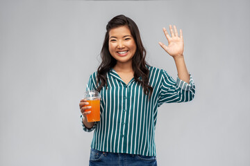 takeaway drinks and people concept - happy smiling young asian woman drinking orange juice from plastic cup with paper straw waving hand over grey background