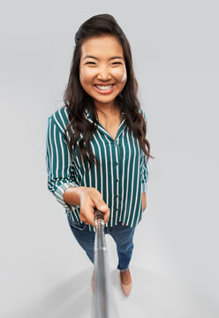 technology and people concept - happy asian woman taking picture with selfie stick over grey background